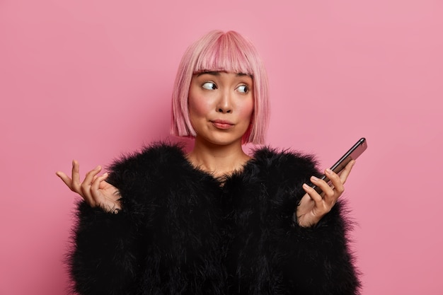 Free photo photo of clueless young woman with pink bob hair looks aside and feels hesitant shrugs shoulders holds mobile phone doesnt know at what time meeting happen wears warm black fluffy sweater poses indoor