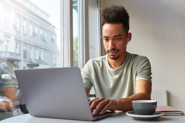 免费照片的照片集中年轻迷人的黑皮肤的男孩,在一台笔记本电脑工作在一个咖啡馆,喝咖啡,若有所思地看着监视器。