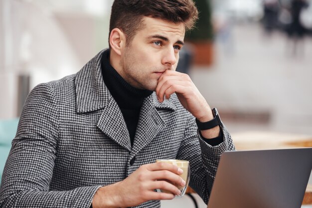 Photo of concentrated businesslike man working with silver laptop in cafe outside, drinking coffee in glass
