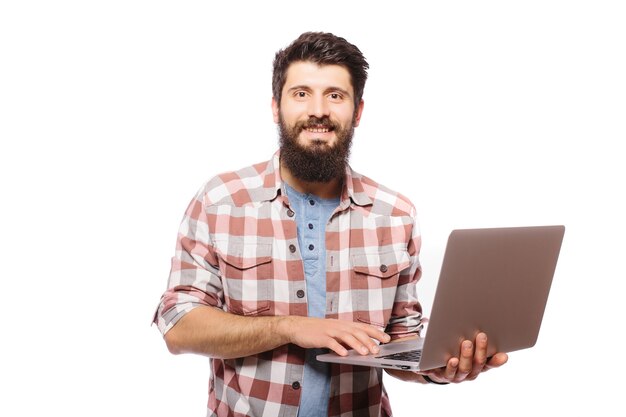 Фото сконцентрированного молодого бородатого человека в очках, одетого в рубашку, с помощью ноутбука, изолированного над белой стеной.