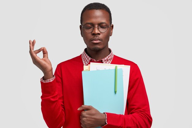 Photo of concentrated young black man meditates indoor, makes ok gesture, carries notepad with pen