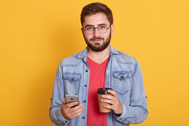 Photo of confident good looking young man holding paper cup with coffee and smartphone, wearing denim jacket, eyewear and t shirt