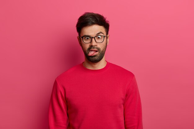 Photo of crazy bearded man sticks out tongue, crosses eyes, feels tired and bored, wears glasses and red sweater, foolishes around, poses against pink wall. Comic face expressions concept