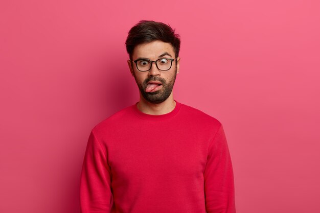 미친 수염 난 남자의 사진은 혀를 내밀고, 눈을 교차하고, 피곤하고 지루하며, 안경과 빨간 스웨터를 입고, 어리석은 짓을하고, 분홍색 벽에 포즈를 취합니다. 만화 얼굴 표정 개념