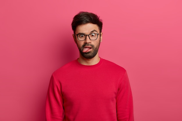 Photo of crazy bearded man sticks out tongue, crosses eyes, feels tired and bored, wears glasses and red sweater, foolishes around, poses against pink wall. Comic face expressions concept