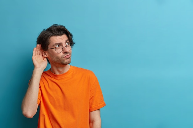Photo of curious man keeps hand near ear and listens private information, tries to overhear gossiping, has intrigued expression, wears round glasses and orange t shirt, copy space on blue wall