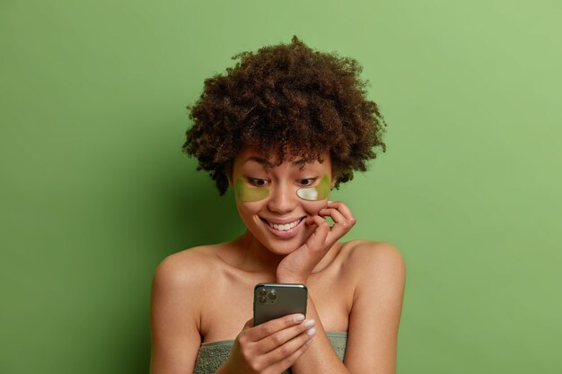 곱슬 머리 만족스러운 여성의 사진은 눈 밑에 녹색 보습 패치를 적용하여 미용 치료를받습니다.