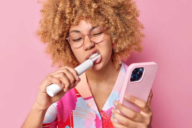 Foto gratuita la foto di una donna dai capelli ricci si lava i denti con una spazzola elettrica usa il dentifricio fa foto di se stessa tramite smartphone isolato su sfondo rosa concetto di cure odontoiatriche e routine di igiene quotidiana