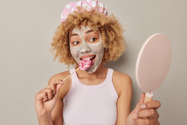 Photo of curly haired woman looks at herself in mirror bites lollipop applies beauty facial mask for rejuvenation dressed in casual t shirt and bathhat isolated over grey background Skin care concept