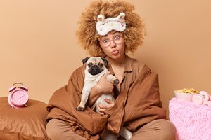 Free photo photo of curly haired young european woman wrapped in soft blanket poses with pug dog in comfortable bed surrounded by pillows alarm clock isolated over beige background domestic atmosphere