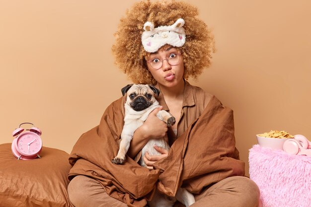 Photo of curly haired young European woman wrapped in soft blanket poses with pug dog in comfortable bed surrounded by pillows alarm clock isolated over beige background Domestic atmosphere