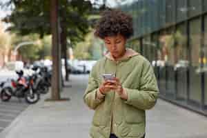 Free photo photo of curly haired young woman in jacket uses mobile phone for chatting online strolls outdoors against blurred urban background holds modern device downloads application for finding route
