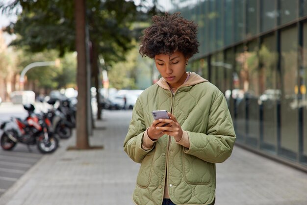 ジャケットを着た縮れ毛の若い女性の写真は、携帯電話を使用して、ぼやけた都市の背景に対して屋外でオンライン散歩をチャットするために、ルートを見つけるための最新のデバイスダウンロードアプリケーションを保持しています