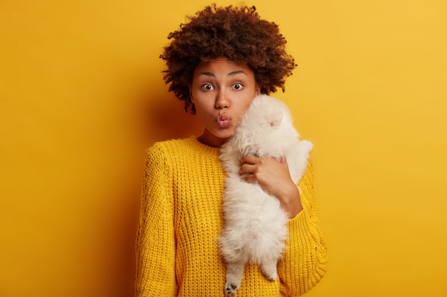 Photo of curly lady carries small white puppy, embraces dog with love, expresses care, prepare for groomer visit, dressed in yellow jumper, stands indoor.