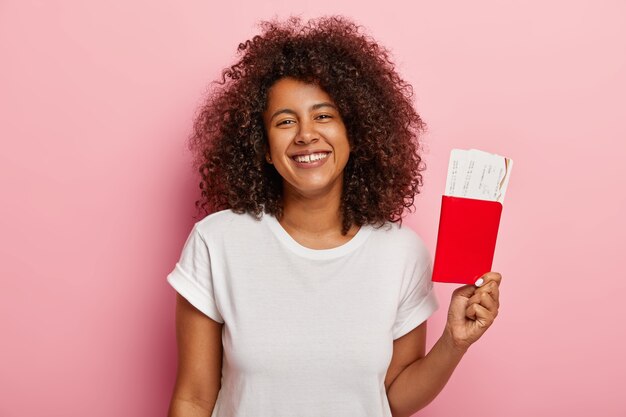 Photo of cute dark skinned woman holds tickets and passport, rejoices summer vacation and trip, happy her dream finally came true, dressed in white t shirt, waits for plane. Traveling concept