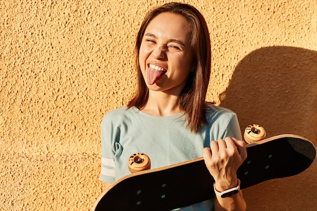 Photo of dark haired cheerful funny woman wear casual t shirt holding skateboard in hands, showing tongue out, posing outdoor isolated over yellow wall.