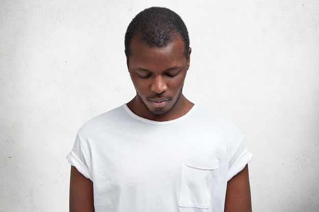 Free photo photo of dark skinned male looks down as notices something, dressed in casual t shirt, advertizes new outfit, isolated over concrete wall.