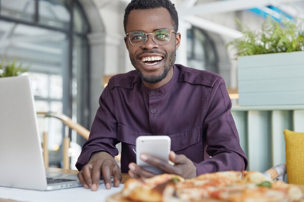 Photo of dark skinned prosperous office manager uses modern cellular for sending messages, works on portable laptop computer