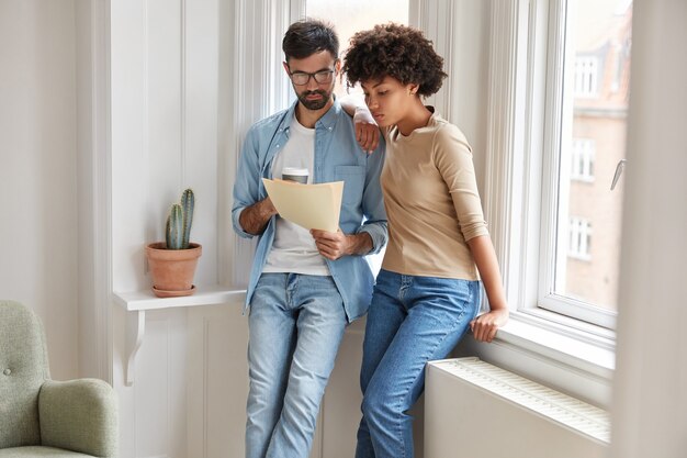 Photo of dark skinned woman and her husband think how satisfy debt, look at bills near the window