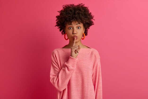 Photo of dark skinned woman presses index finger to lips, demands keep silence, shushing mysteriously, prepares secret for someone, makes hush gesture, dressed casually, isolated on pink wall