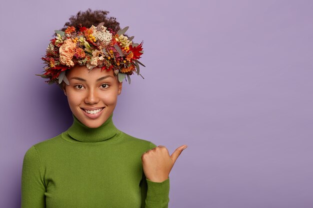 Photo of dark skinned young curly woman points thumb away, wears green turtleneck, autumnal wreath, has pleasant smile, demonstrates copy space for your advertising content, gives suggestion