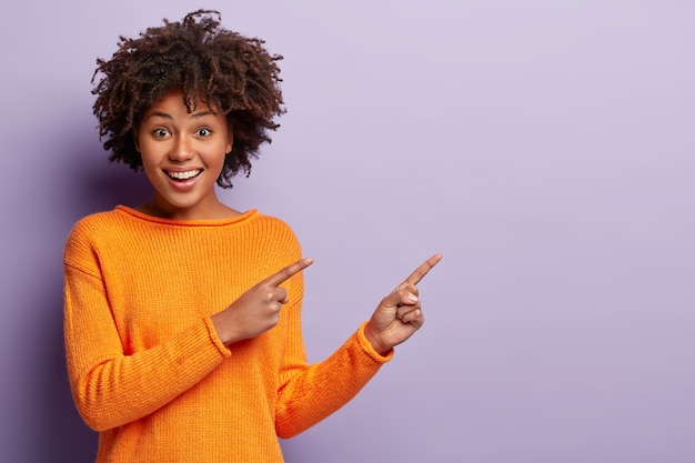 Photo of delighted African American woman points away with both index fingers, promots awesome place for your advertising content