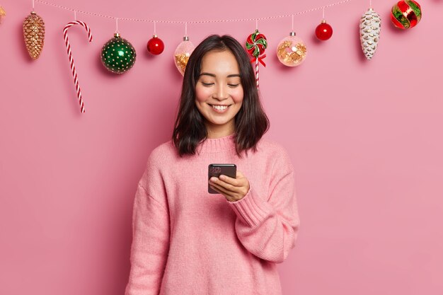 Photo of delighted brunette Asian woman with eastern appearance holds modern mobile phone sends messages of congratulation on New Years Eve wears casual jumper poses 