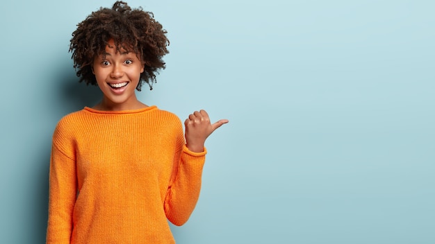 Photo of delighted cheerful Afro American woman with crisp hair, points away, shows blank space, happy to advertise item on sale, wears orange jumper, demonstrates where clothes shop situated