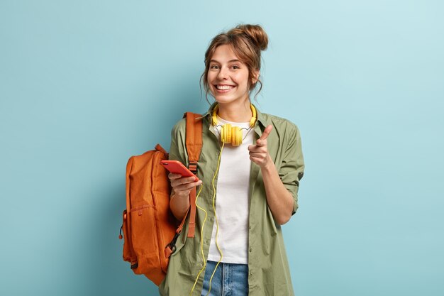 Photo of delighted European woman points at you happily, holds smartphone device connected to headphones, uses music app