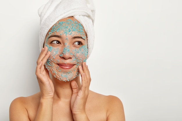 Photo of delighted female model stands topless against white wall, touches face skin, makes peeling with sea salt scrub, removes pores and white spots. Skin solution and spa treatment concept