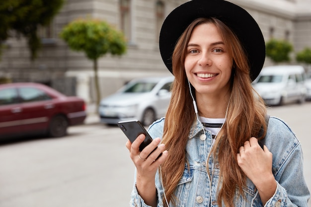 Photo of delighted female model with European appearance, smiles gently at camera, listens song from radio website or audiobook