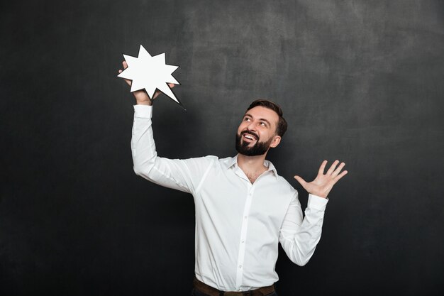 Photo of delighted man holding blank speech star and looking aside over dark gray wall, copy space