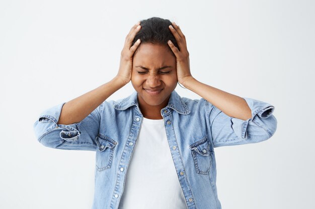 Photo of disappointed Afro-american woman in denim shirt holding her hands on temples frowning face and closed eyes in despair and terror. Woman regreting her act. Female in despair and shock