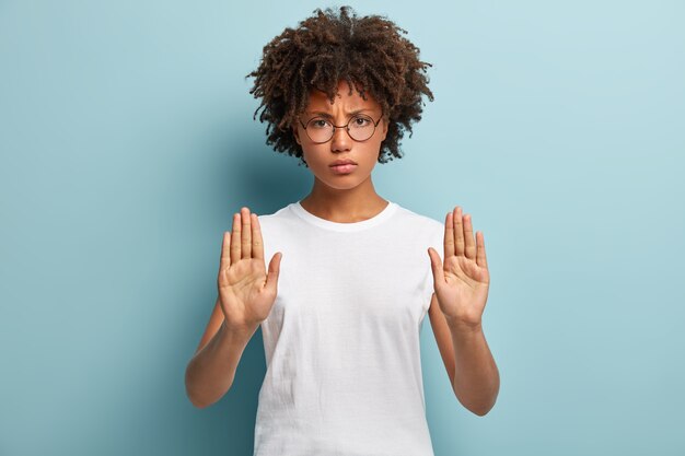 Photo of discontent dark skinned female with angry facial expression, shows palms, asks to stop talking, refuses doing something, wears transparent glasses and casual t shirt, models indoor