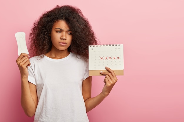 Photo of discontent lovely curly haired woman looks at calendar with marked pms days