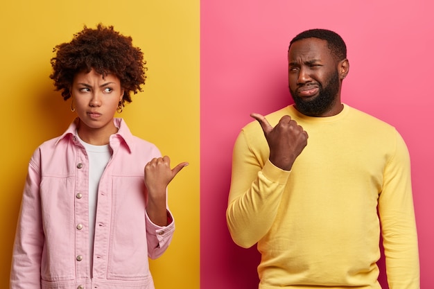 Photo of discontent pessimistic ethnic curly girlfriend and boyfriend point thumbs at each other, smirk faces unhappily