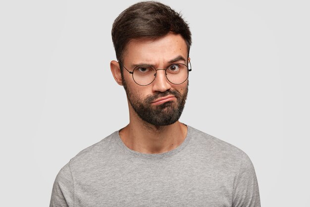 Photo of discontent unshaven young male frowns face and purses lips, has displeased expression, wears grey t-shirt, raises eyebrows, models against white wall. People and emotions concept