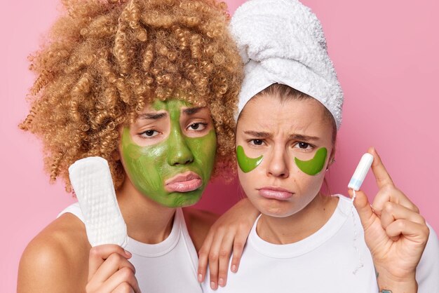 Photo of discontent young women look sadly at camera apply green beauty mask and hydrogel patches under eyes for skin treatment hold sanitary napkin and tampon feel unhappy isolated over pink wall