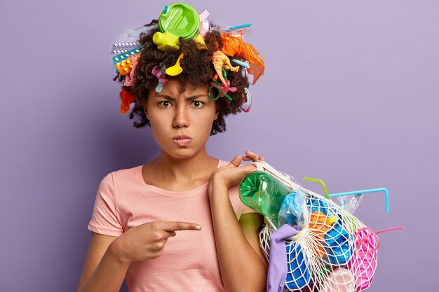Photo of displeased Afro American woman angry with abusive use of plastic, points at bag with collected garbage, has wastes in head, isolated on purple wall. Non recyclable pollution concept