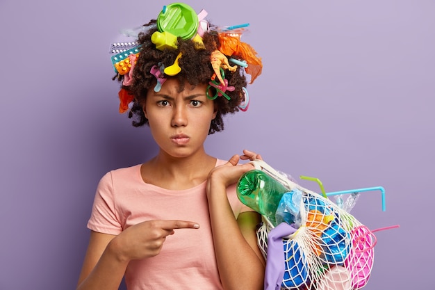 プラスチックの乱用に腹を立てている不機嫌なアフリカ系アメリカ人女性の写真、収集されたゴミの入ったバッグを指差して、頭にゴミがあり、紫色の壁に隔離されています。リサイクル不可能な汚染の概念