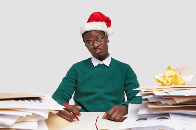 Photo of displeased black man feels apathy as doesnt want to work, dressed in festive headgear and sweater, surrounded with piles of documents