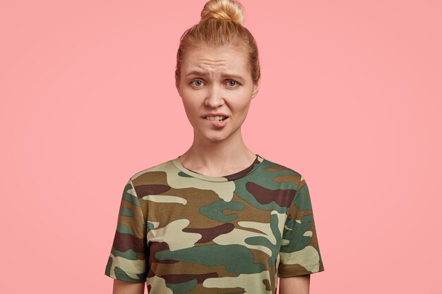 Photo of displeased blonde female model looks with negative emotions, bites lower lip, raises eyebrows, feels concerned and dissatisfied, wears casual t shirt, isolated over pink wall