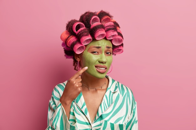 Photo of displeased ethnic woman points at problematic zone on face, indicates at cheek and shows pimple, wears green moisturizing face mask, applies hair rollers, wears casual dressing gown. Beauty