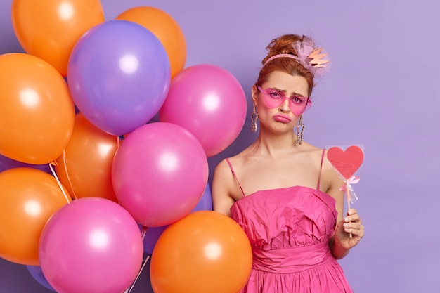 Free photo photo of displeased redhead young woman looks with sullen expression at camera holds delicious candy multicolored balloons poses indoor against purple background