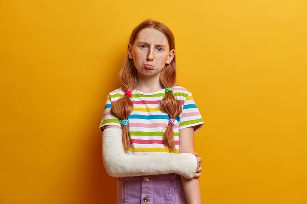 Photo of displeased small preteen girl has bad mood, purses lips and looks in discontent, being offended by close friend, has hurt feelings, dressed casually, poses with broken arm