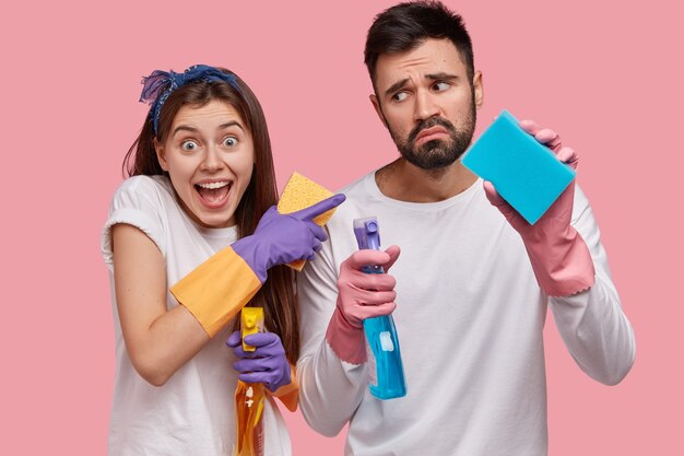 Photo of displeased unshaven man holds detergent and sponge, looks in dissatisfaction