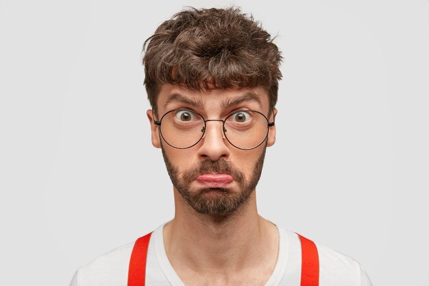 Photo of displeased unshaven man purses lower lip, has dark hair, looks in bewilderment at camera