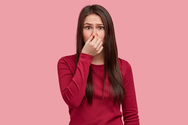 Photo of displeased woman closes nose with reek, feels terrible smell from garbage, wears red clothes