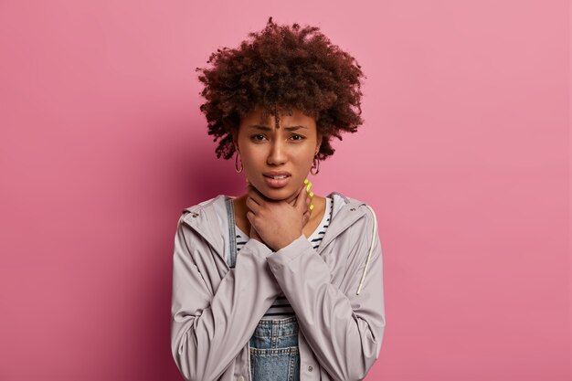 Photo of displeased woman grabs neck with both hands, feels chocking, asks to bring pills from sore throat, stands dissatisfied from feeling discomfort, gets ill, wears anorak, poses over rosy wall