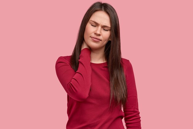 Photo of displeased woman keeps hand on neck, eyes shut, has long hair, wears red sweater